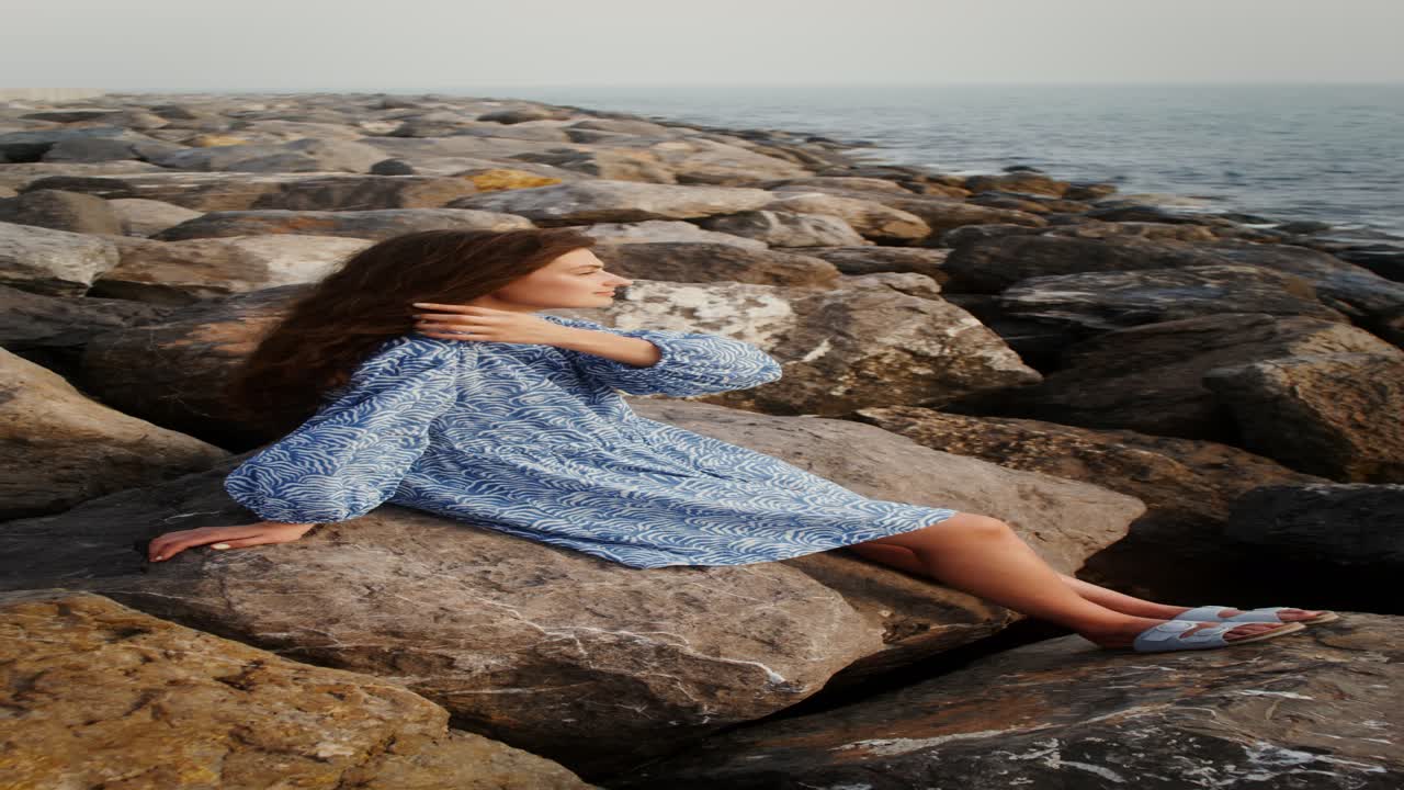 женщина в синем платье сидит на скалах у моря
