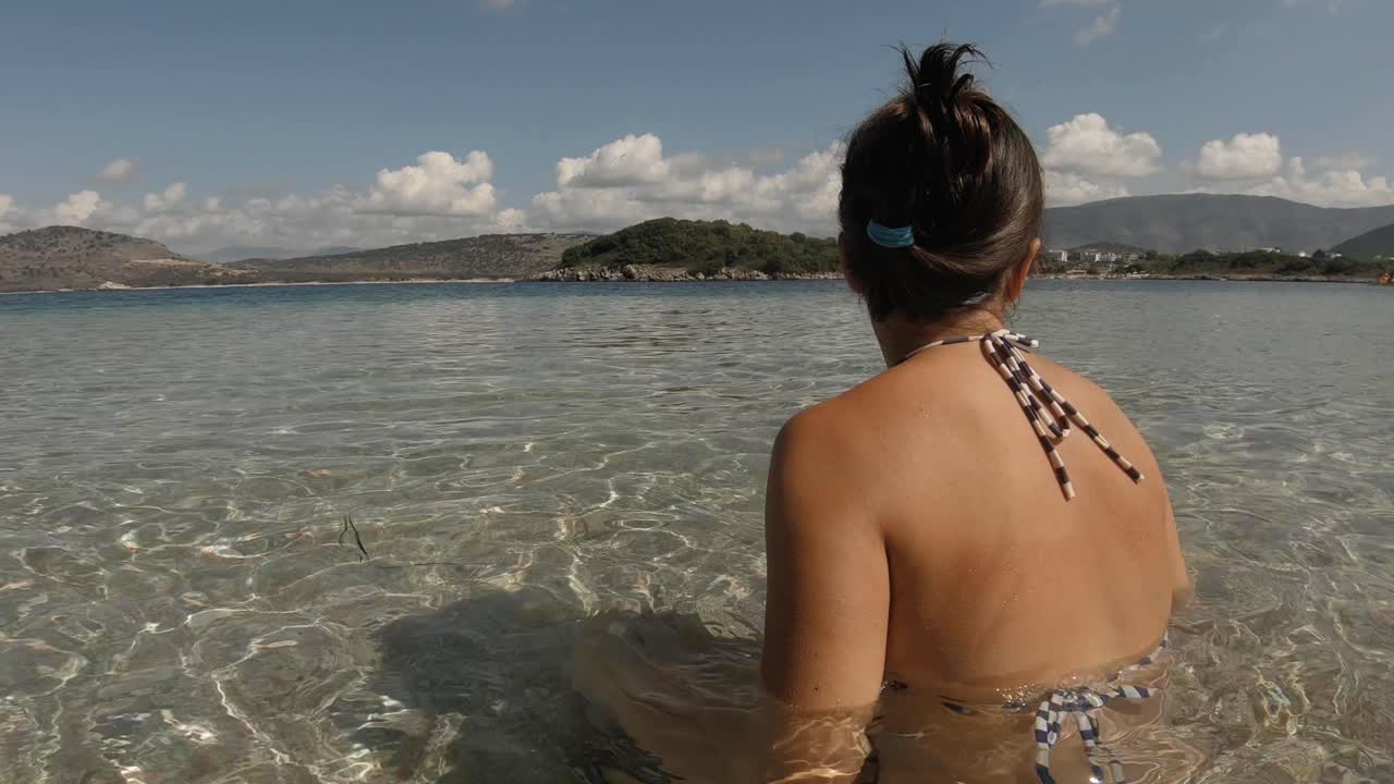 Back view of a girl siting on a beach looking in the horizon. Clear warm water aound her.