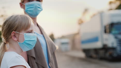 woman with a child in protective masks are standing near a dirty dusty road in the city