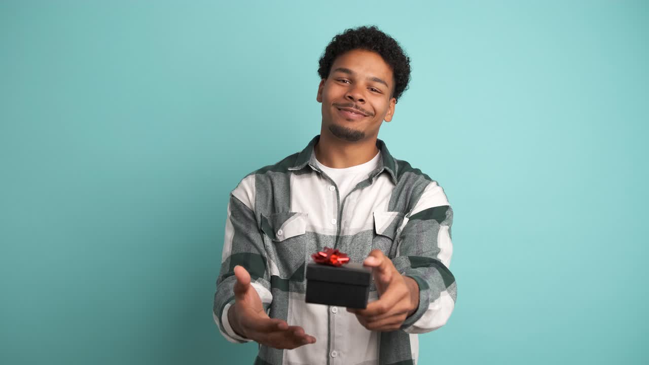 Happy man giving gift box and looking at camera in blue studio