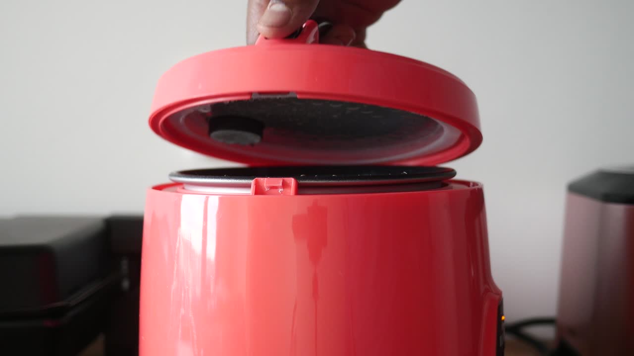 A person opening a pink mini rice cooker lid, followed by a view of the open rice cooker