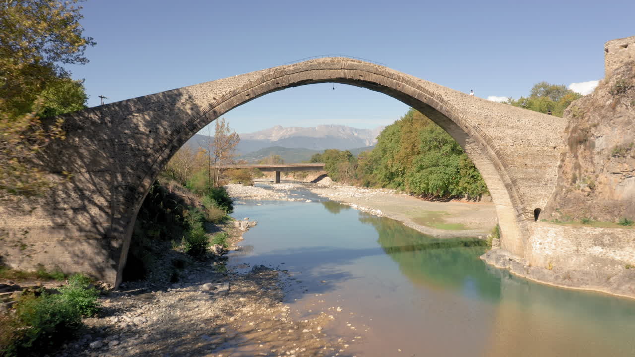 Zagori, Yunanistan'da sonbahar ormanıyla çevrili dağ nehri üzerindeki taş kemerli Konitsa köprüsü, orta irtifa drone çekimi.