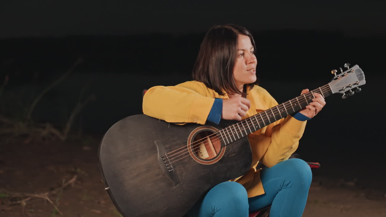 Donna sola in campeggio di notte che suona la chitarra, strimpellando lenti accordi acustici accanto a braci incandescenti, indossando una giacca gialla su una sedia pieghevole, accordando e componendo sotto il cielo stellato della foresta, un momento intimo e riflessivo.