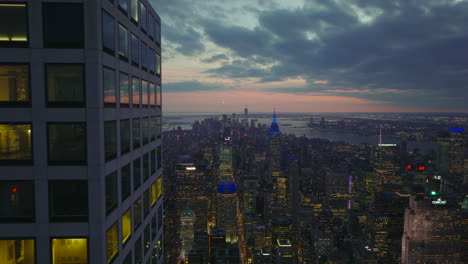 Tight-fly-above-top-storeys-of-modern-skyscraper-towering-above-city.-Reveal-panoramic-shot-of-downtown-at-dusk.-Manhattan,-New-York-City,-USA