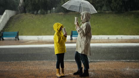 Happy-teenage-girl-in-a-yellow-jacket-drinks-water-and-communicates-with-her-mom-while-standing-under-an-umbrella-in-the-park-during-heavy-rain