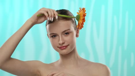 young woman holding gerbera flower with gentle smile a young woman with a gentle smile holds a vibrant orange gerbera flower against a serene blue background