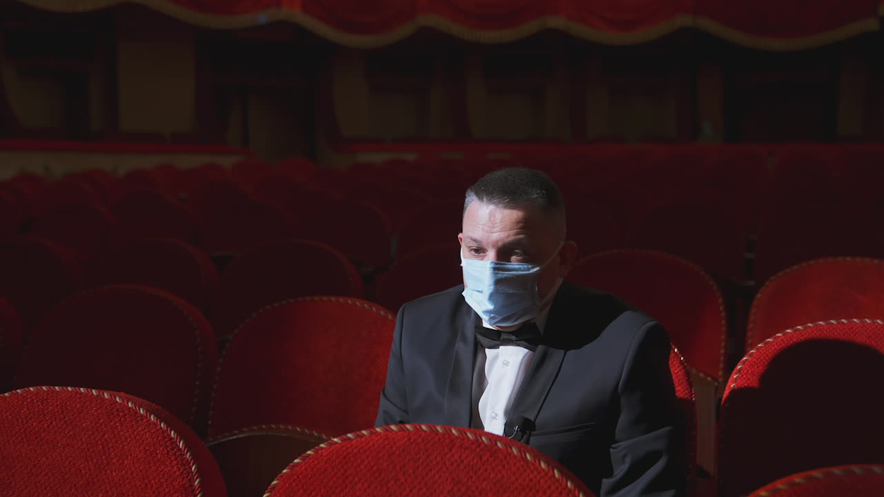 Sad man in medical mask in empty theater. Rich man in suit sitting alone among empty red armchairs and sleeps in opera hall. Lockdown concept.