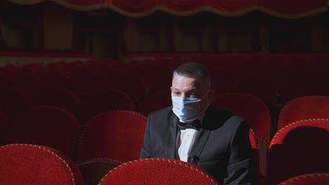 sad man in medical mask in empty theater. rich man in suit sitting alone among empty red armchairs and sleeps in opera hall. lockdown concept.