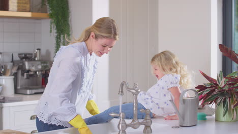 mother wearing rubber gloves at home in kitchen with young daughter having fun and washing girl's feet as they do washing up at sink- shot in slow motion