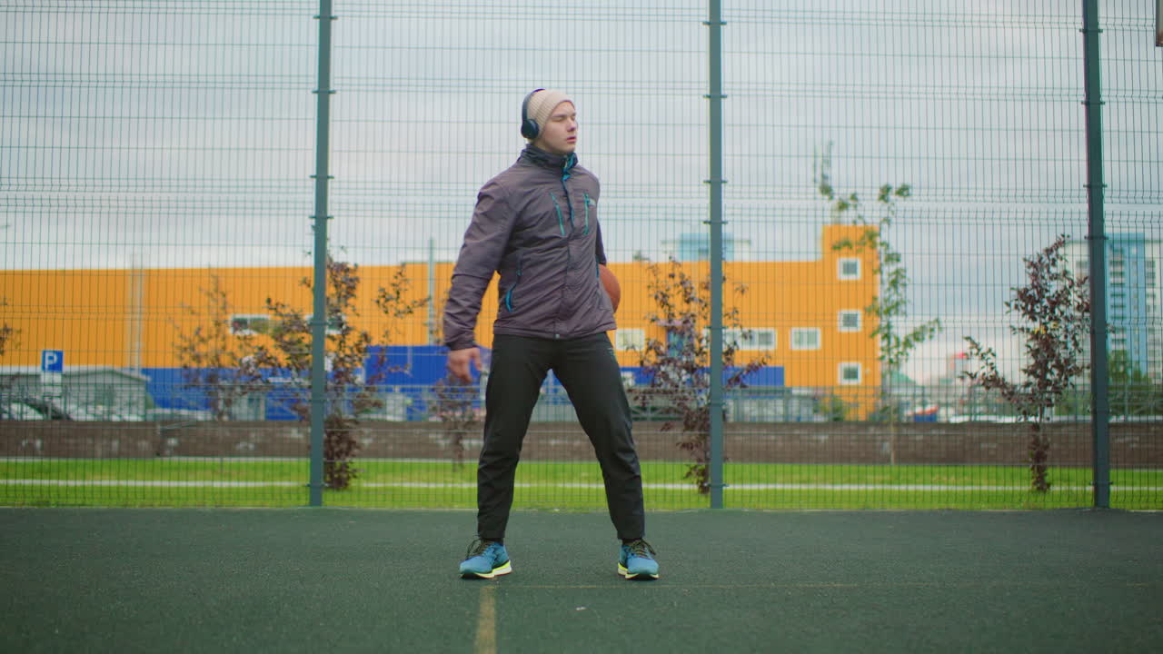 White man on urban basketball court wearing jacket and beanie, headphones on, holding basketball and warming up overcast sky, fenced turf, industrial skyline in background, solo street athlete