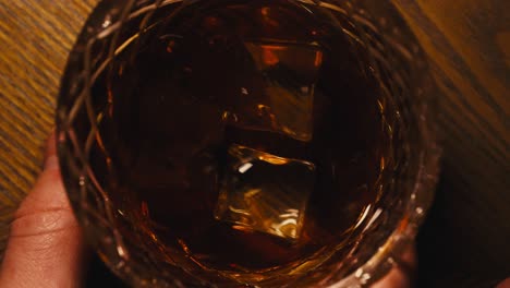 close up top down slow motion shot of a man swirling a glass of bourbon whiskey on a wooden table