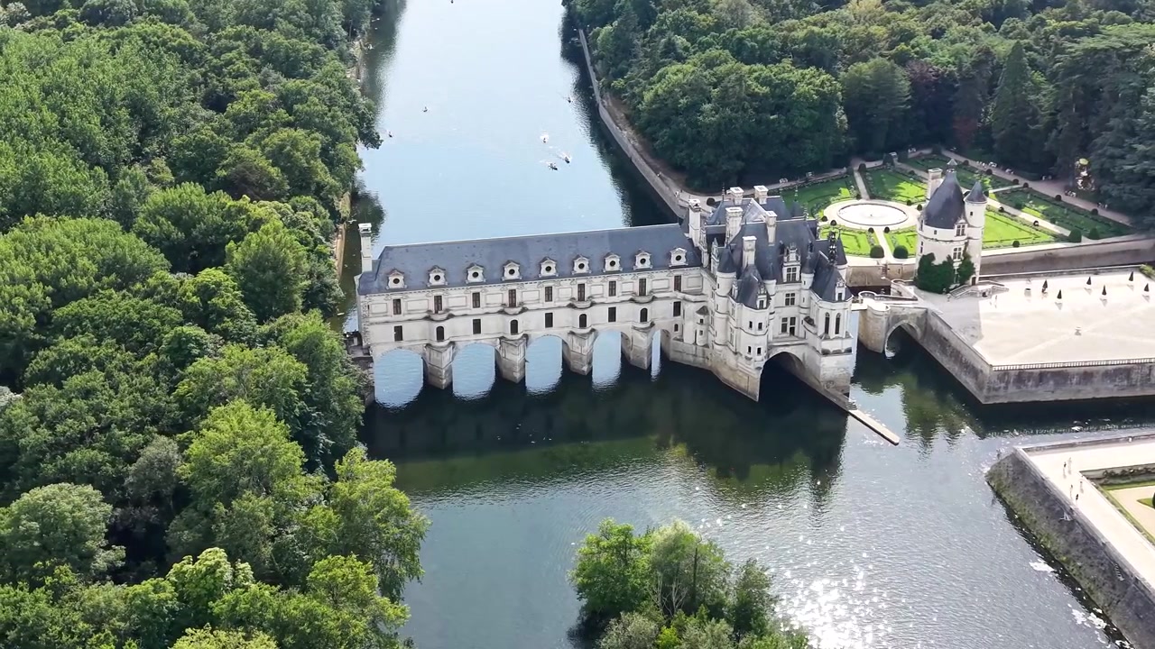Vue aérienne cinématographique par drone du Château de Chenonceau enjambant le Cher, mettant en valeur ses arches au-dessus de l'eau, son architecture Renaissance et ses jardins à la française.
