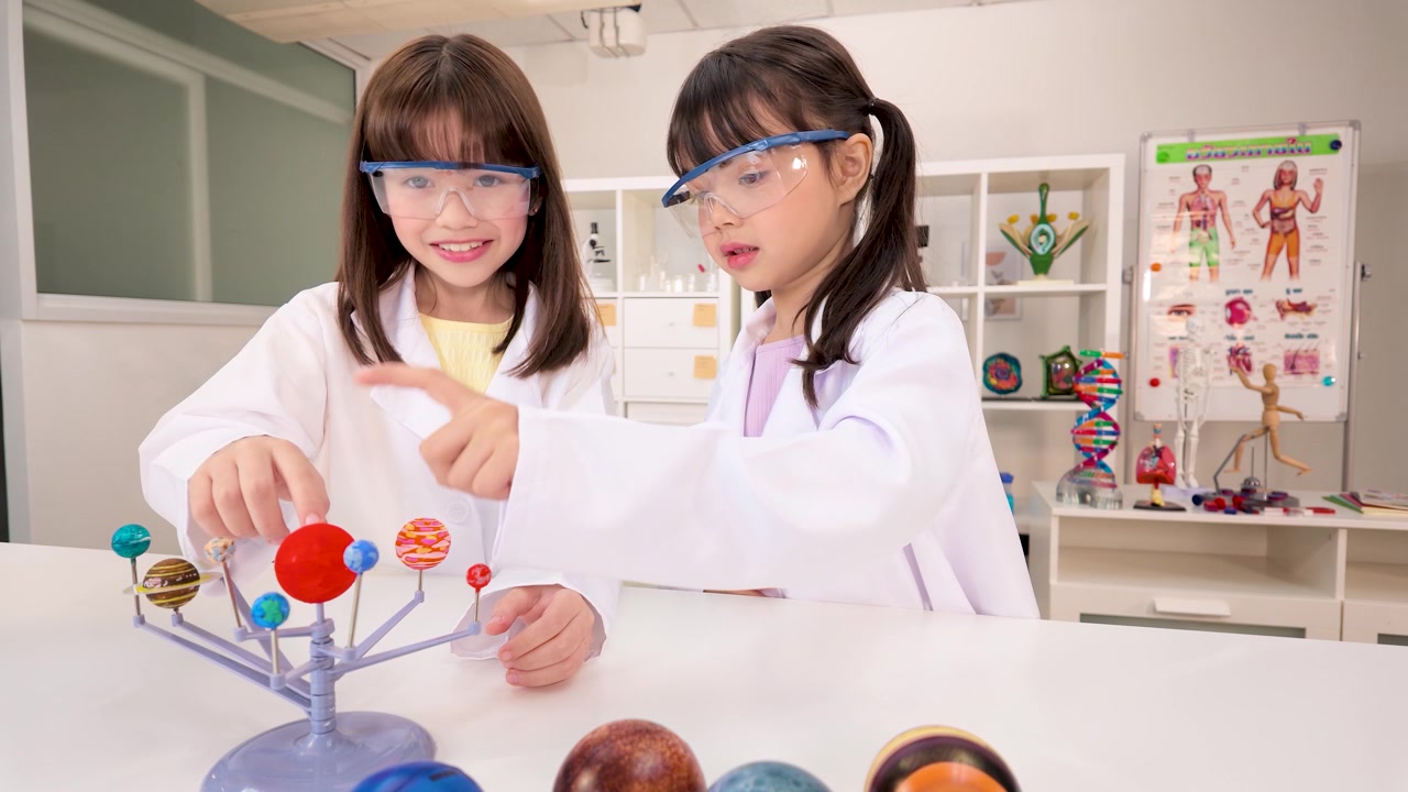 Two young girls in lab coats and goggles examine a solar system model in school.
