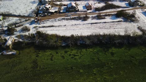 Drohnenaufnahme-Nach-Einem-Van,-Der-An-Einem-Schönen-Wintertag-Entlang-Des-Flusses-Cetina-Fährt-Und-An-Schneebedeckten-Ländlichen-Häusern-Vorbeifährt,-Dalmatiner,-Kroatien