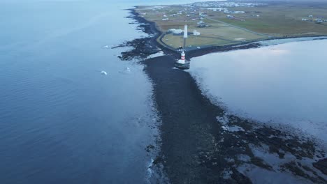 Drone-Volando-Lejos-Del-Faro-En-La-Costa-Islandesa-Y-Revelando-Una-Hermosa-Playa-Cubierta-De-Arena-Negra