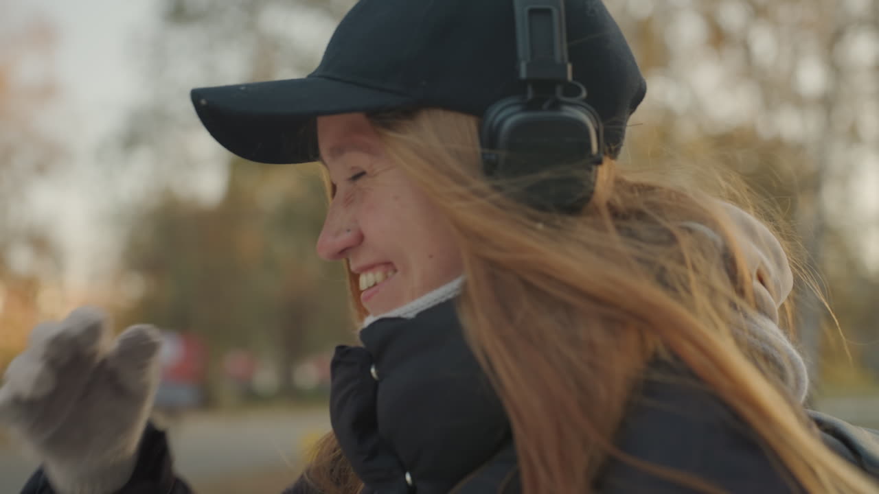 Donna caucasica sorridente con le cuffie che si gode la brezza del parco autunnale, indossa un berretto nero, una giacca imbottita e guanti di lana, capelli rossi scompigliati dal vento e risate allegre creano un'atmosfera calda e spontanea, i cambi di inquadratura suggeriscono