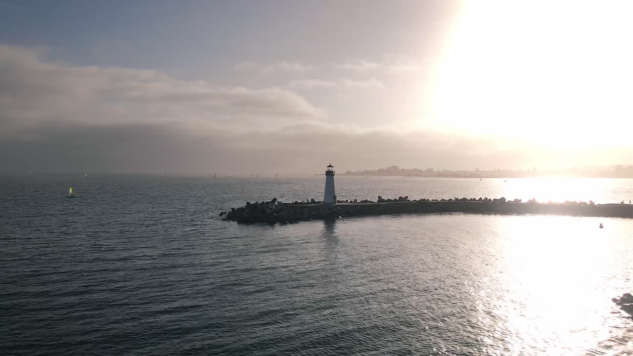 A slow motion drone shot panning left in front Walton Lighthouse and the small crafts harbor in Santa Cruz California with the sun setting in the background with sailboats in the Pacific Ocean