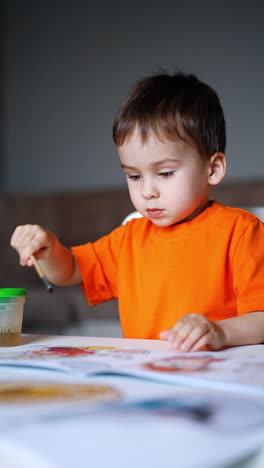 lovely kid holding a brush in the cup. baby boy pulls the sheet of paper book closer to color it. vertical video.