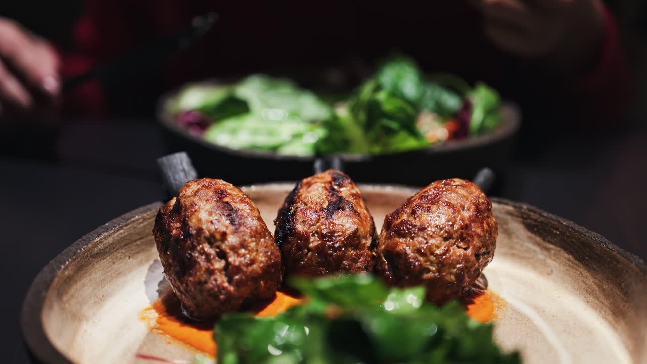 Three grilled meat skewers on charcoal sticks served on a plate with an orange sauce with a woman cutting and eating a salad at a restaurant on the background
