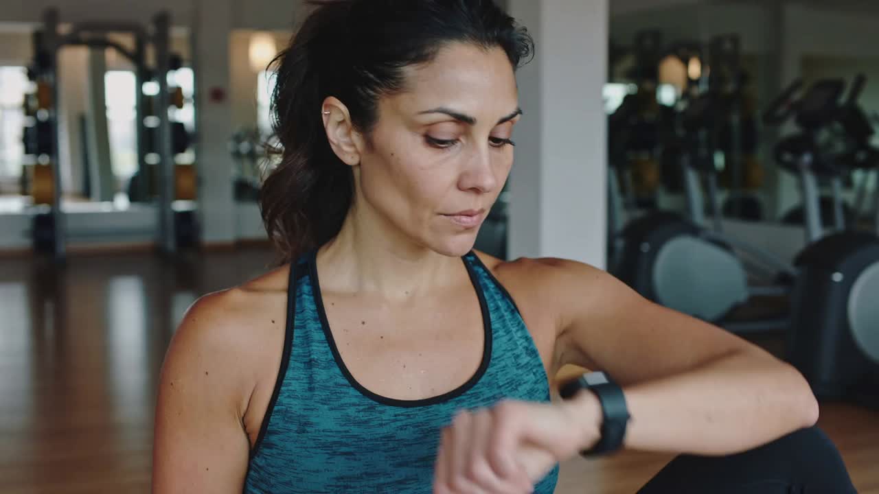 Looking at smartwatch, woman in teal top reading, tapping in gym, checking stats before resuming