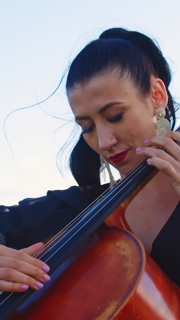 Dark-haired female musician uses no bow to play violoncello. Woman moving her head in the rhythm of music. Mountains backdrop in blur. Vertical video
