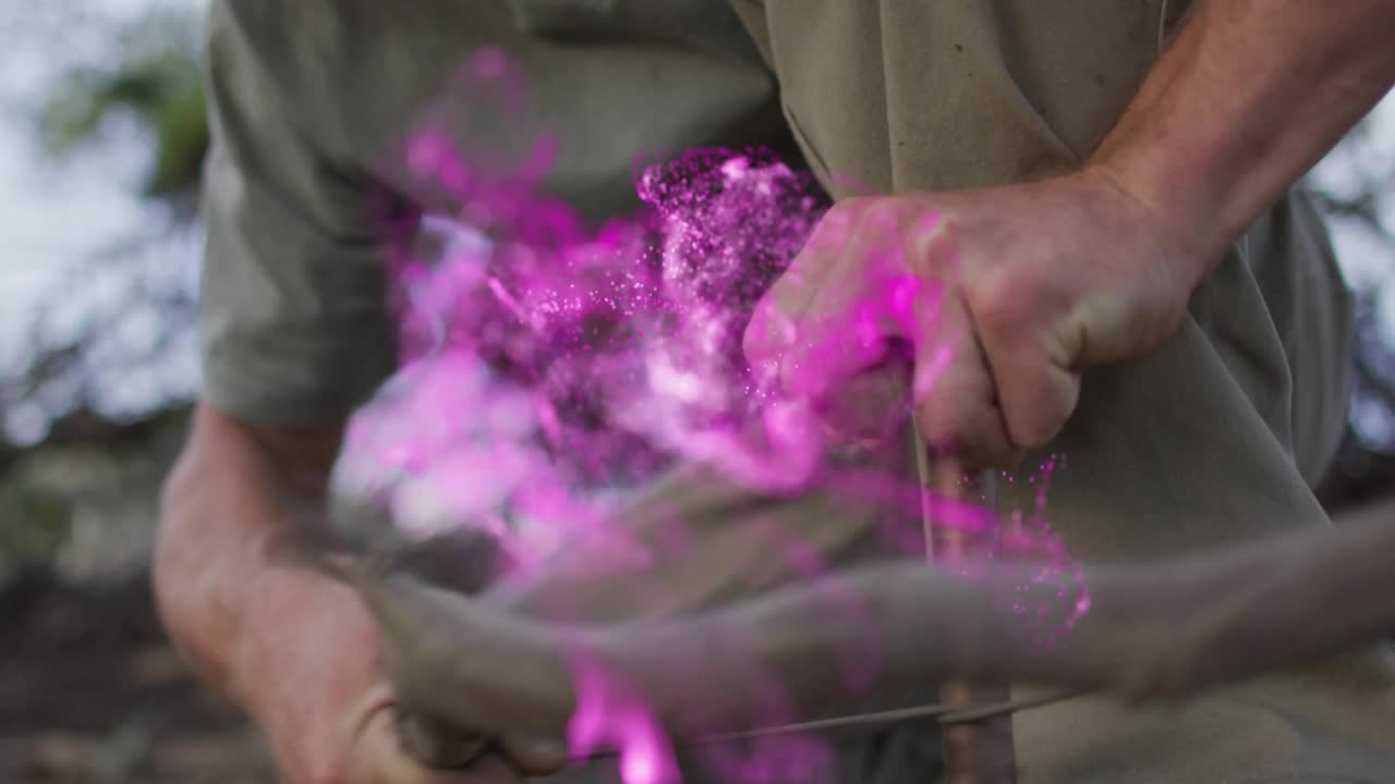 Un uomo inginocchiato in una radura boschiva usa un trapano ad arco per la sopravvivenza, creando scintille con fumo viola.