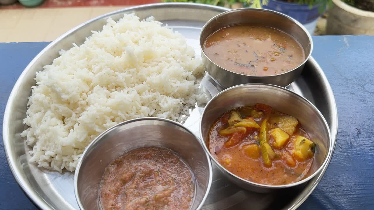 a traditional Indian meal served on a stainless steel plate, The meal comprises steaming hot white rice served with three small stainless steel bowls containing dal, vegetable curry, and chutney