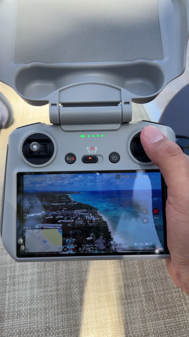 Close-up vertical shot of a person holding a drone remote controller with a built-in screen displaying live aerial footage of a tropical beach and coastline