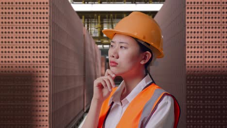 close up side view of asian female engineer with safety helmet thinking and looking around then raising her index finger while standing with red brick packed in stacks are stored