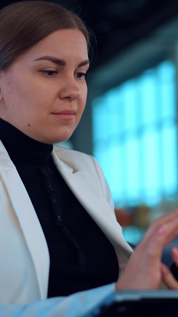Business lady wearing white jacket sits in comfortable armchair focused on her phone. Caucasian woman working using her smartphone. Close up. Blurred backdrop. Vertical video