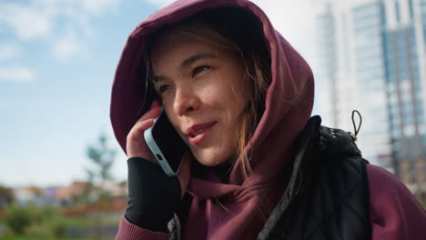 young lady in mauve hoodie and black jacket chats on phone while beaming under bright sunny sky in bustling urban courtyard bordered by modern high rises, capturing candid tech lifestyle moment