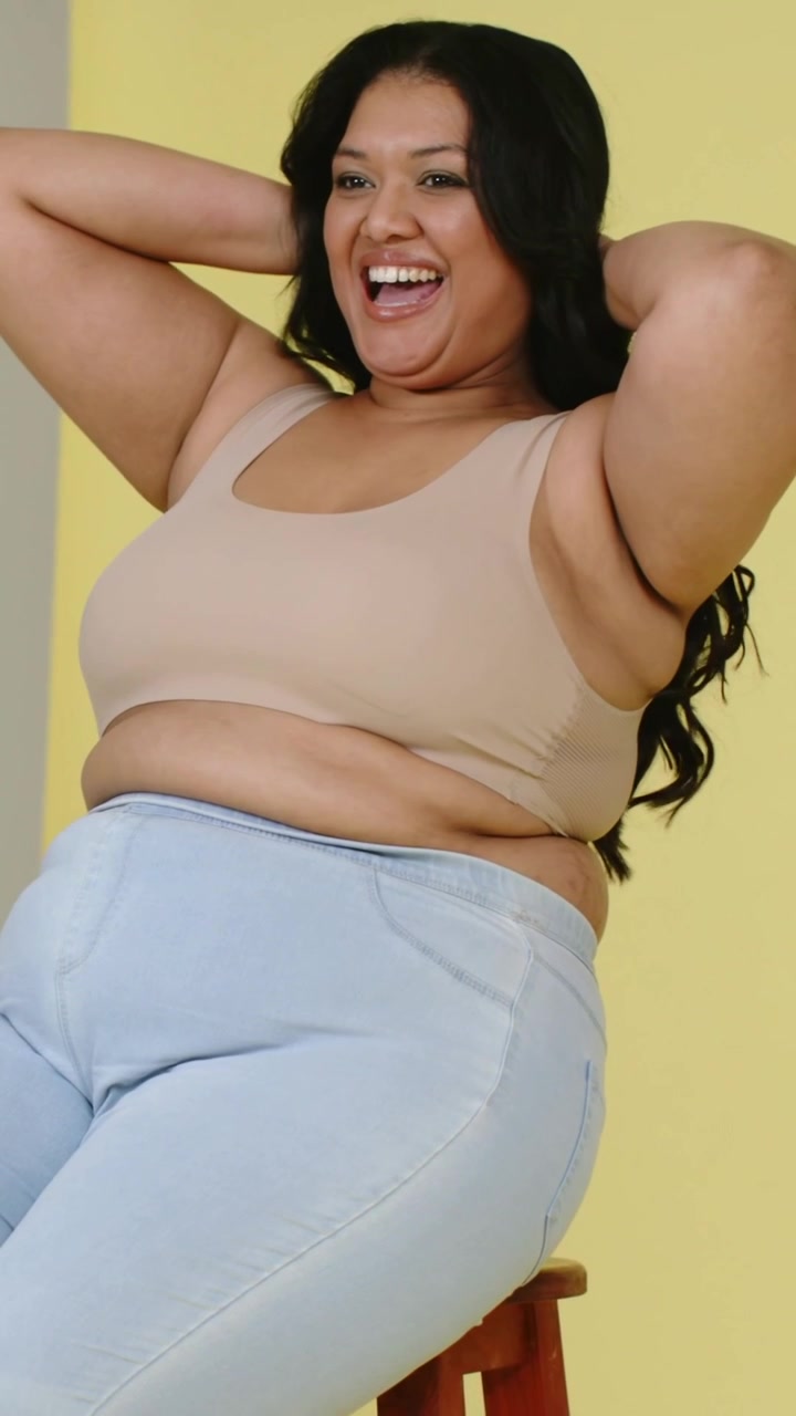 Video verticale: Donna sorridente si sistema i capelli su uno sgabello in studio, il fotografo la dirige, indossa un top beige e jeans.