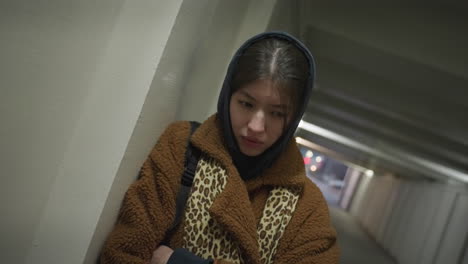 갈색 코트를 입은 소녀가 지하 통로의 벽에 기대어 깊은 우울증을 보이고 있습니다. 엄숙한 표정과 철수 된 자세는 희미하게 밝은 곳에서 그녀의 감정적 고통을 강조합니다.