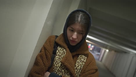 una chica con un abrigo marrón se apoya contra la pared de un túnel subterráneo, mirando profundamente deprimida. expresión solemne y postura retirada, destacando su angustia emocional en un dimamente iluminado