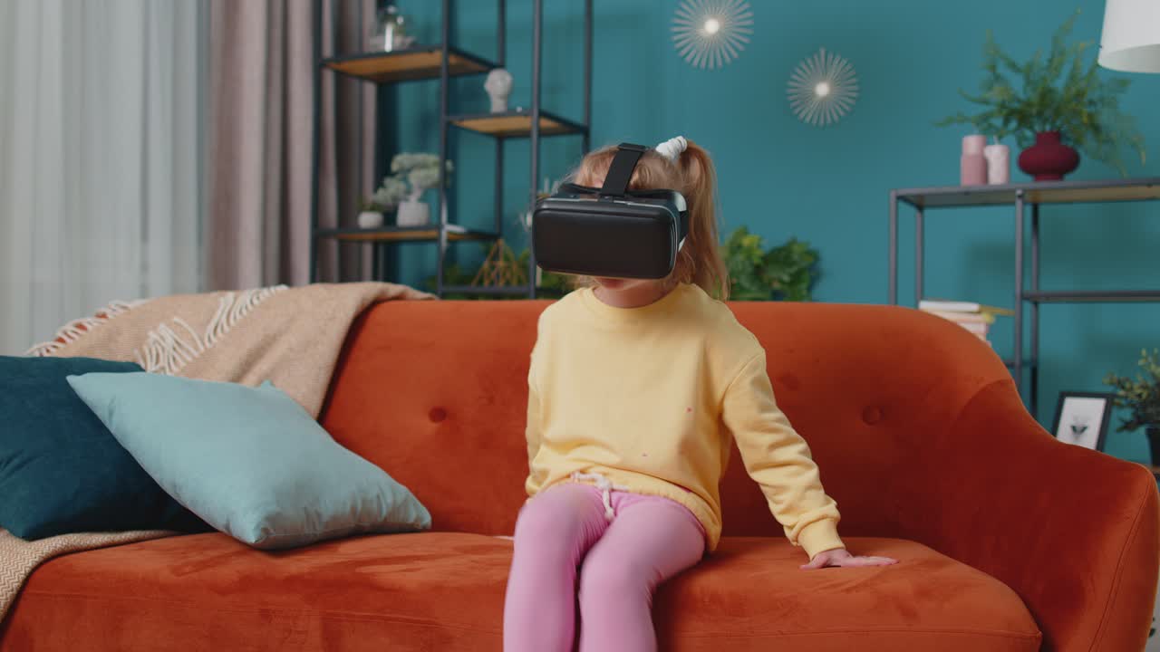 Toddler girl sitting on home sofa using virtual reality headset helmet app to play video 3d game