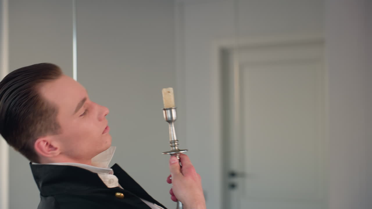 young man dances dramatically reaching up to remove candle from stand, then inspects candle thoughtfully in bright minimalist studio with mirror reflection and elegant coat movement