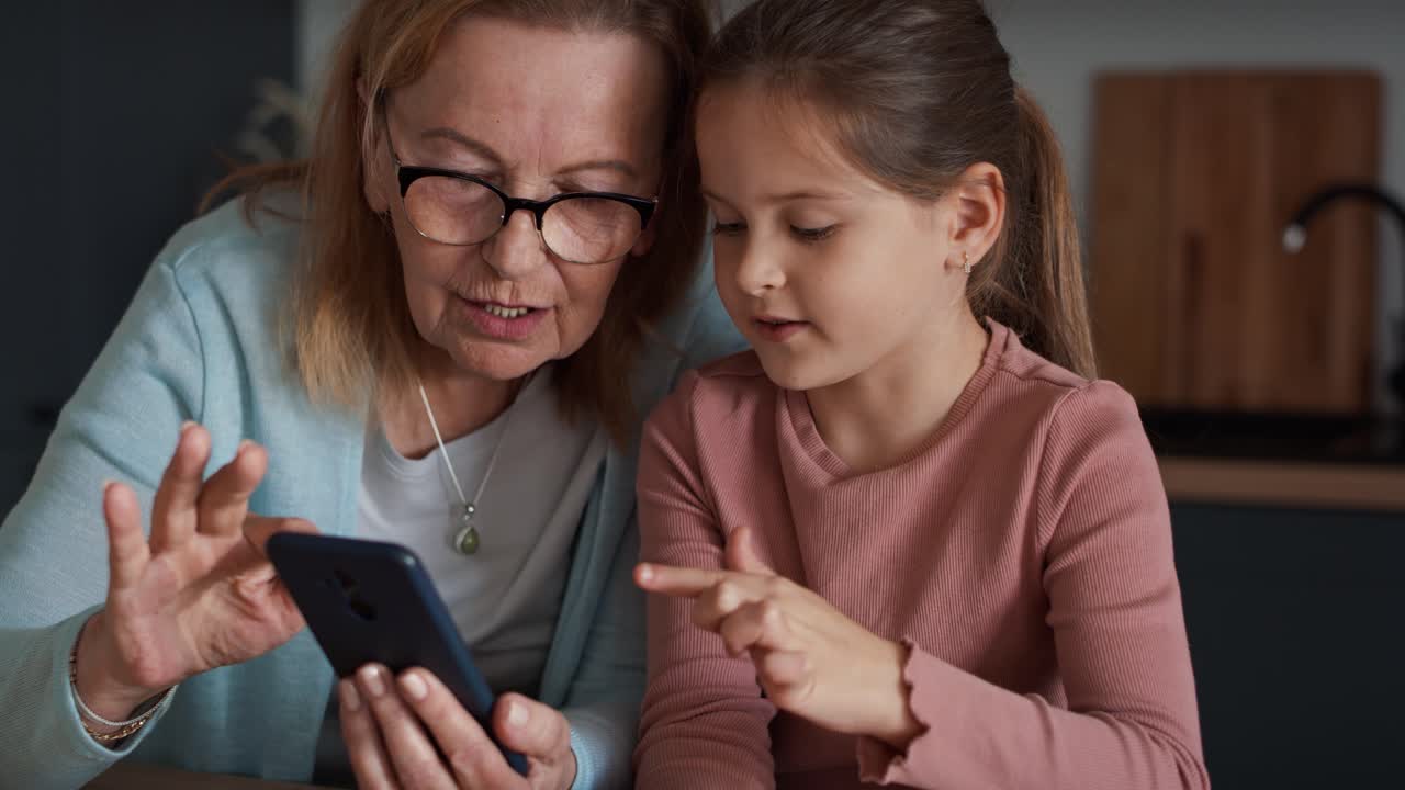 Frontaloptagelse af kaukasisk bedstemor og barnebarn, der bruger mobiltelefon i køkkenet og har det sjovt