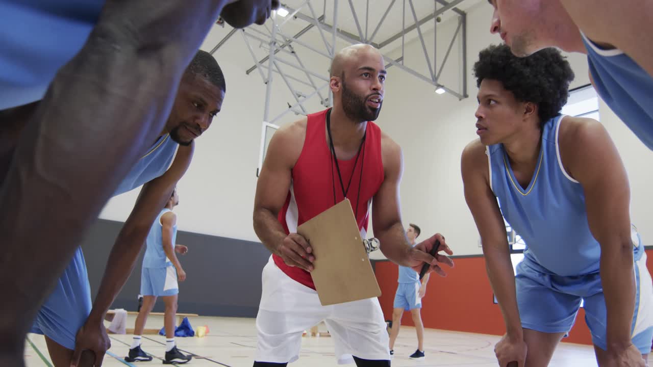 Diverse male basketball team and coach with clipboard discussing in huddle on court, in slow motion