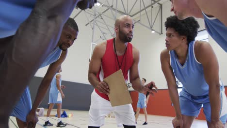 Diverse-male-basketball-team-and-coach-with-clipboard-discussing-in-huddle-on-court,-in-slow-motion