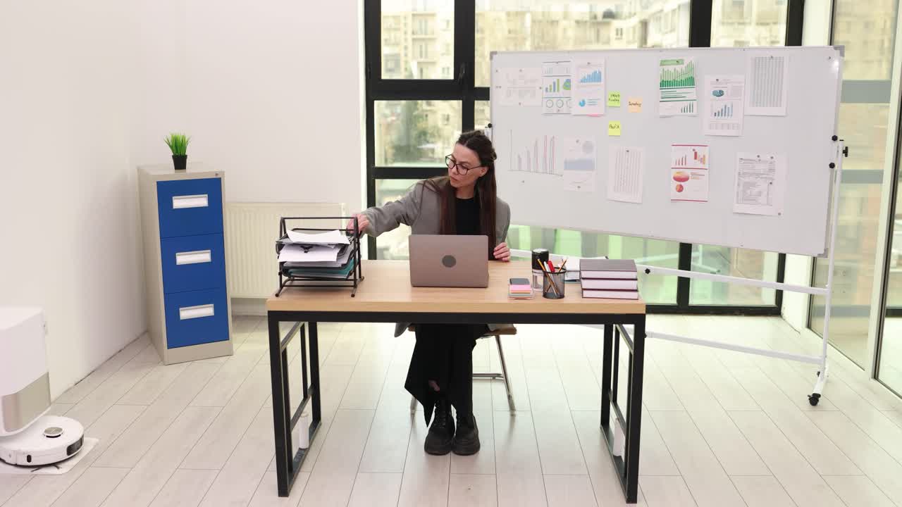 Mujer de oficina revisa documentos consultando la pantalla de su portátil en la mesa.