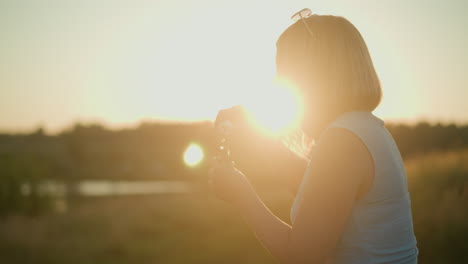 woman in blue sleeveless top with sunglasses on her head blowing bubbles during golden hour, sunlight gently illuminating her hair, creating beautiful rim light, with soft blurred natural background