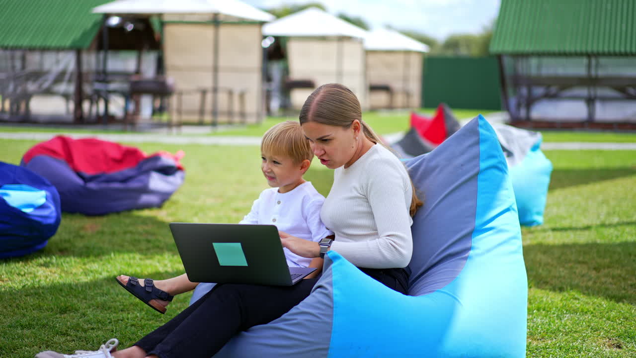 Young woman and her son using laptop at their rest. Mother and her boy watching something and discussing it. Rest zone at backdrop in blur.
