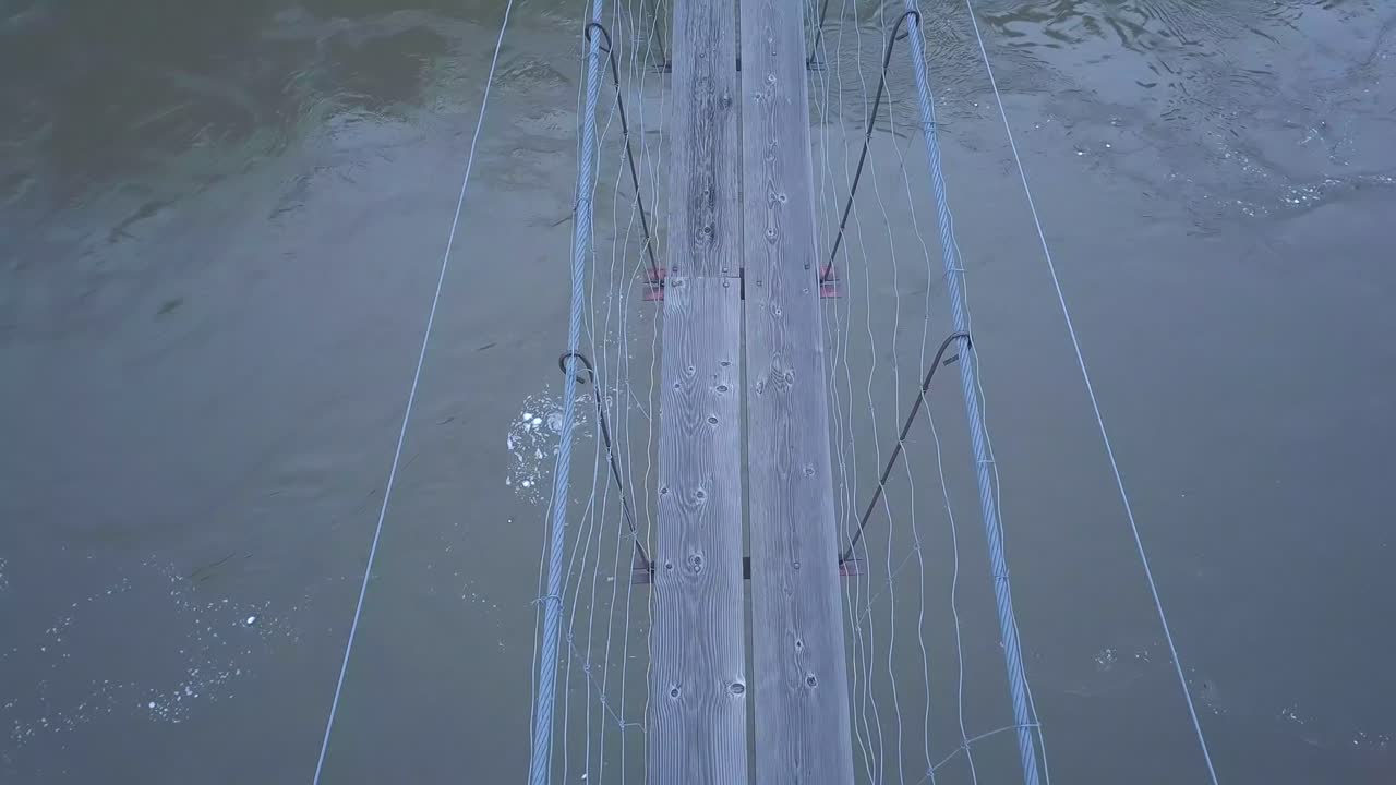 Aerial POV of narrow suspension foot bridge over swirling muddy river