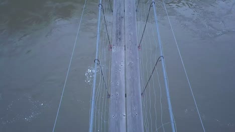aerial pov of narrow suspension foot bridge over swirling muddy river