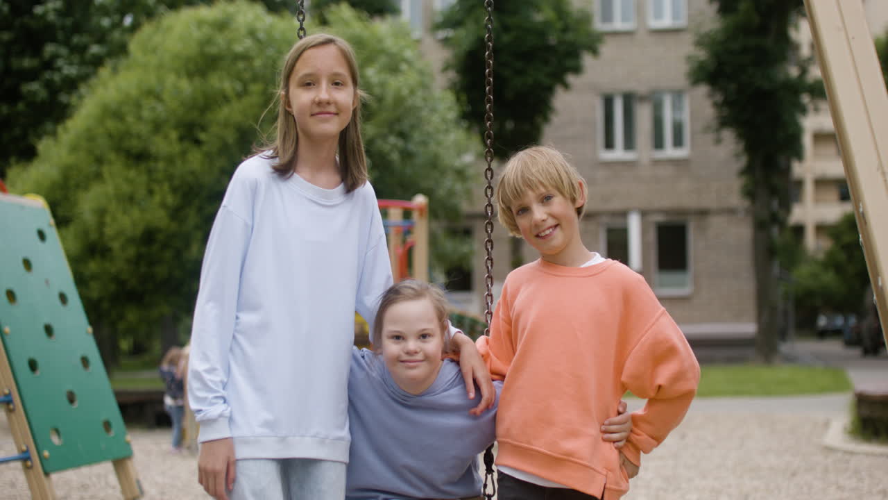 Little girl with down syndrome sitting on a swing and smiling at camera. Her friends are next to her