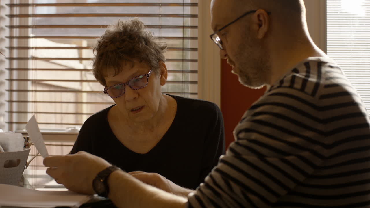 An elderly woman sits at a kitchen table with a younger man. They are sorting, reviewing, and discussing documents together. Both wear reading glasses. Warmly backlit by the sun through window. 24fps.