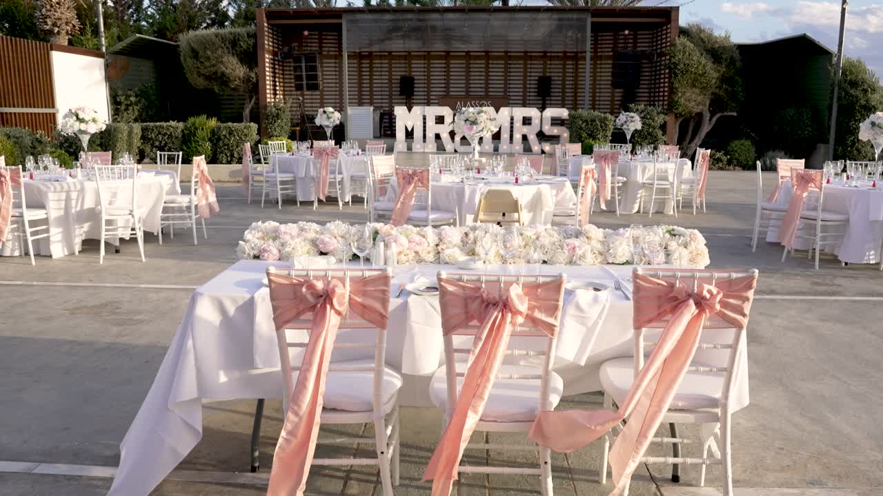 I tavoli sono disposti all'esterno per festeggiare un matrimonio. I tavoli sono coperti da tovaglie bianche. Delle fasce rosa sono legate alle sedie. I fiori sono posizionati sui tavoli e le decorazioni sono disposte intorno all'area.