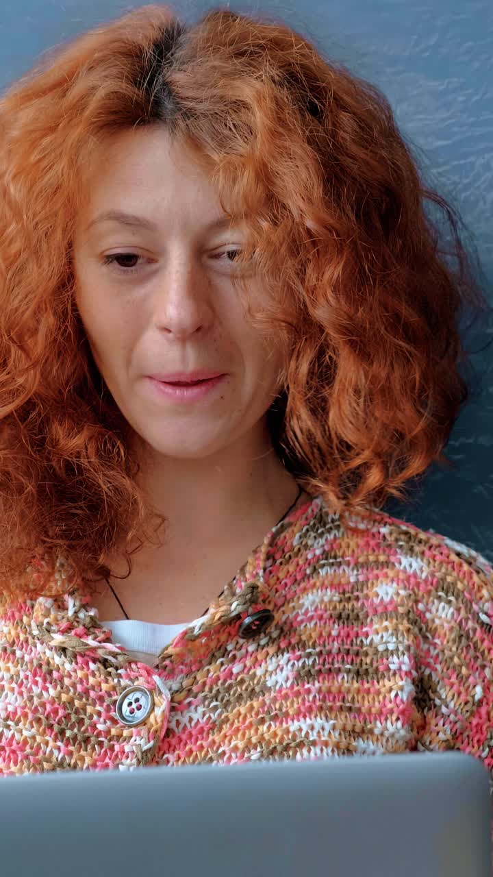 Woman with curly red hair is smiling while using a laptop, showcasing a joyful moment of engagement with technology in a cozy indoor setting