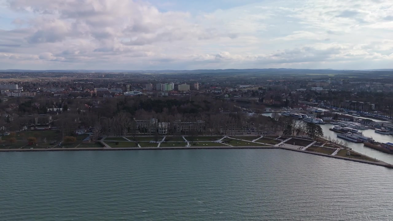 A Siófok sul Lago Balaton, la costa urbana con il suo porto turistico, il molo e gli appartamenti vicini costeggia il lungomare pubblico e la vegetazione in una vista laterale con drone sotto un cielo coperto