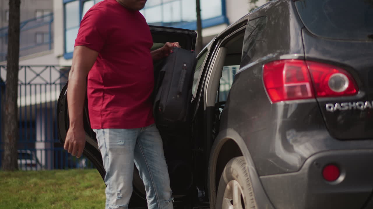 Uomo nero che scende dall'auto con camicia rossa e jeans, utilitaria parcheggiata a bordo strada vicino a un condominio, che controlla la portiera posteriore e lo pneumatico, scena urbana diurna che trasmette partenza di routine e concentrazione.